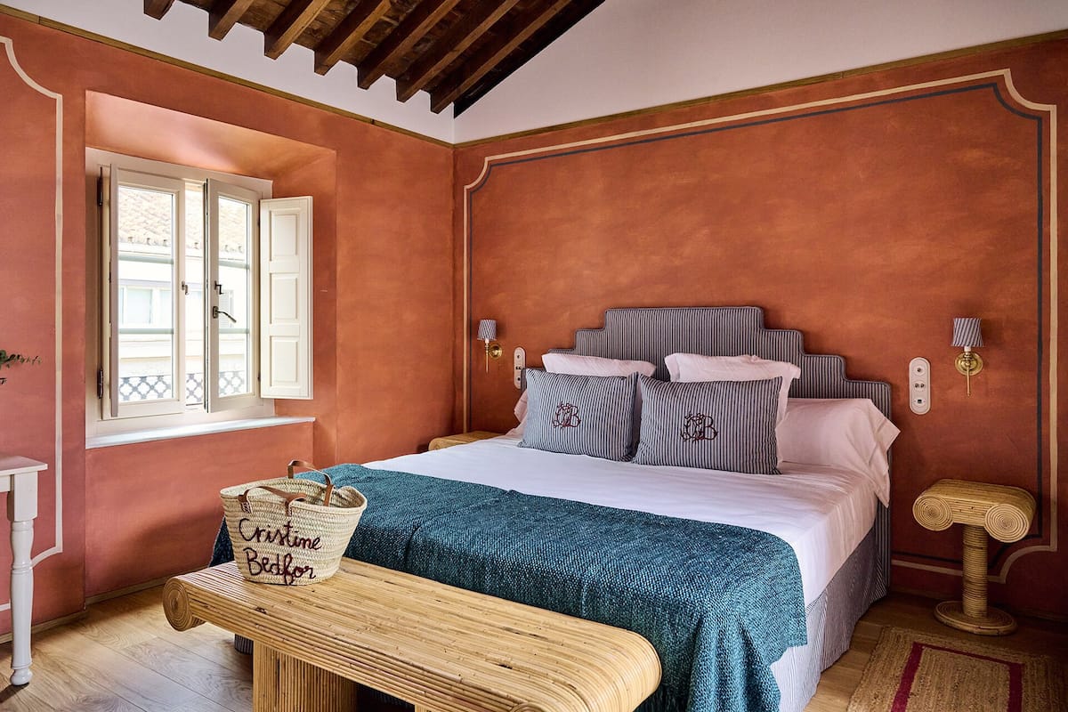 A bedroom with a bed, bedside lamps, a bedside table, a window, and a bench.