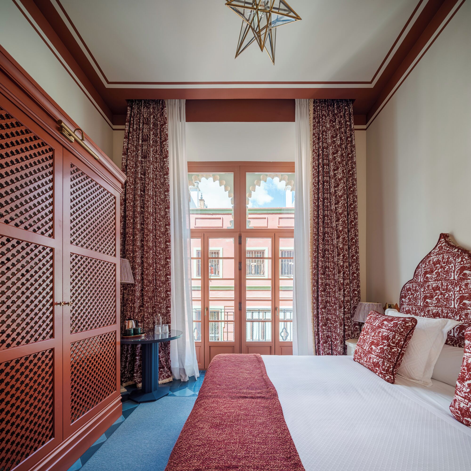 A hotel room with a bed, curtains, a window with a view, a star-shaped ceiling light, and a patterned wardrobe.