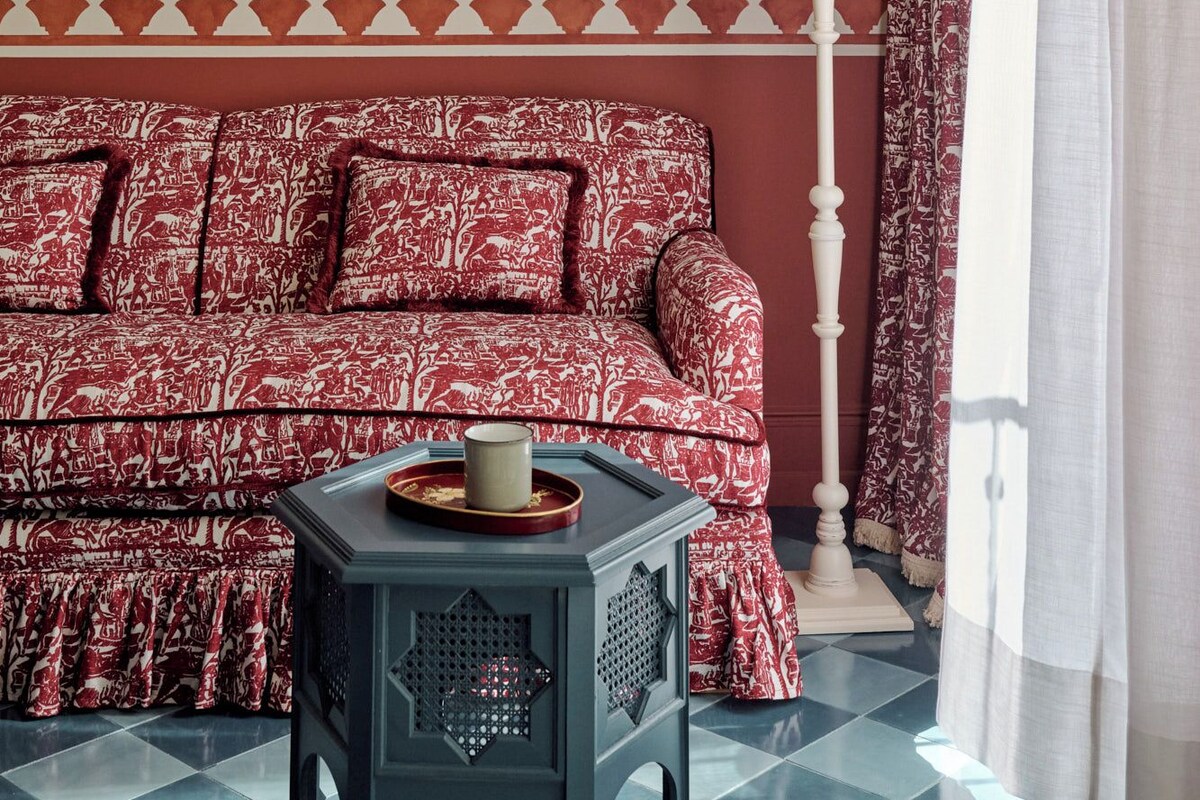 A room with a checkered floor, a sofa with a patterned cover, a small table with a decorative item, a lamp, and curtains.