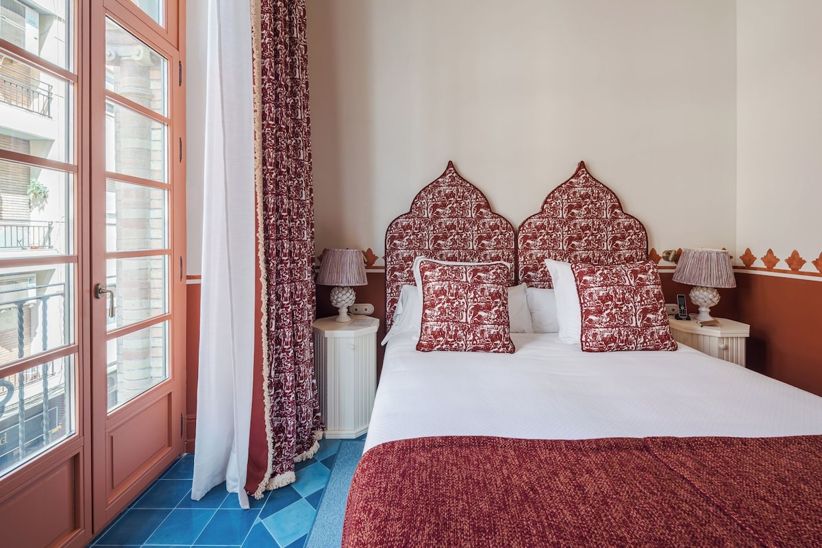A hotel room with a large bed, patterned pillows, a nightstand with lamps, a window with curtains, and a blue-tiled floor.