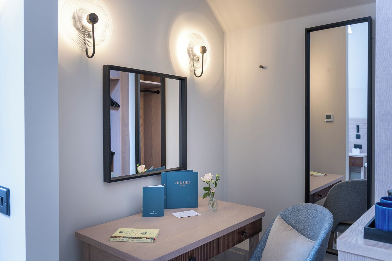 A hotel room with a wooden desk, a mirror, two wall-mounted lights, a vase with flowers, and a large mirror reflecting the room.