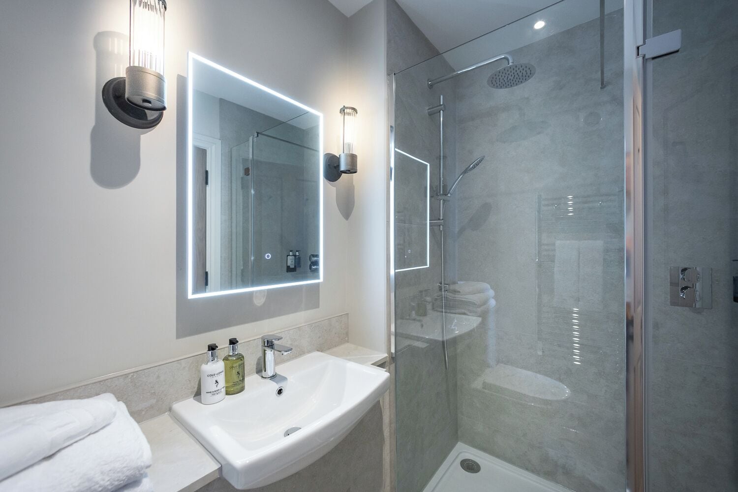 A modern bathroom with a glass-enclosed shower, a large mirror with integrated lighting, and a white sink with toiletries.