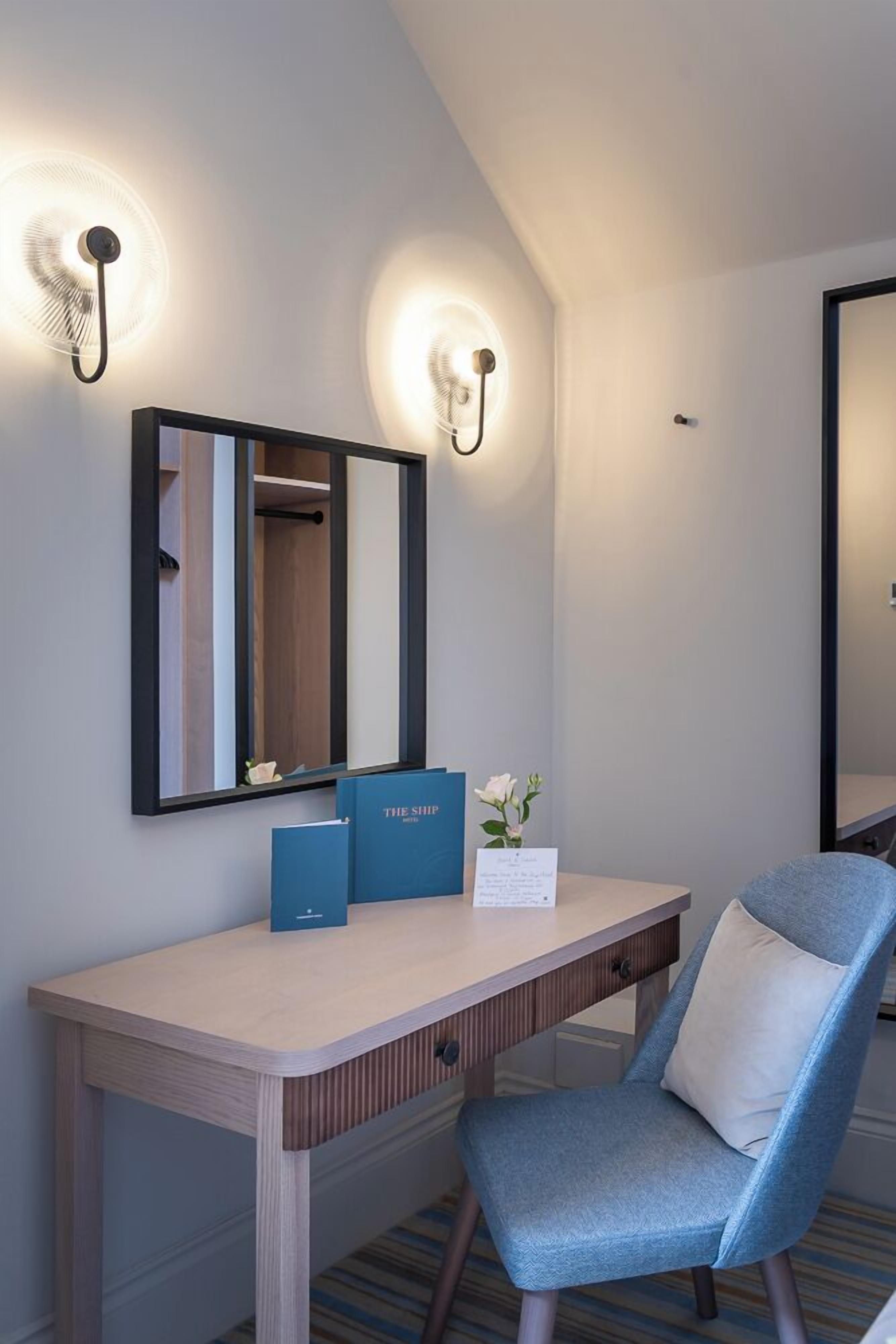 A hotel room with a desk, a chair, a mirror, and wall-mounted lights.
