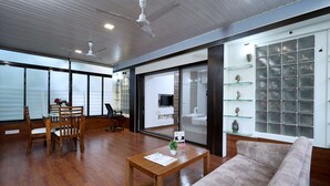 A modern living room with a wooden floor, a sofa, a coffee table, and a dining area with a table and chairs. There is a large window with a view, a television, and a decorative shelf with items.