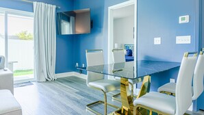 A modern room with a blue wall, a glass table with gold legs, white chairs, and a mounted TV. - 3BR/2.5 Bath Townhome Golf Sim, Theatre by NMB Sport Complex (Little River)