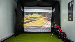 A room with a golf simulation setup, including a screen displaying a golf course, artificial turf, and golf bags. - 3BR/2.5 Bath Townhome Golf Sim, Theatre by NMB Sport Complex (Little River)