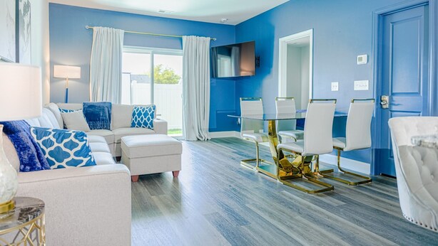 A modern living room with a blue accent wall, a white sofa with patterned cushions, a glass dining table with gold chairs, and a flat-screen TV mounted above the table. - 3BR/2.5 Bath Townhome Golf Sim, Theatre by NMB Sport Complex (Little River)