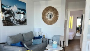 A modern living room with a grey sofa, a round mirror, and a painting of a mountain landscape. There are two small round tables with lamps and a vase of flowers.