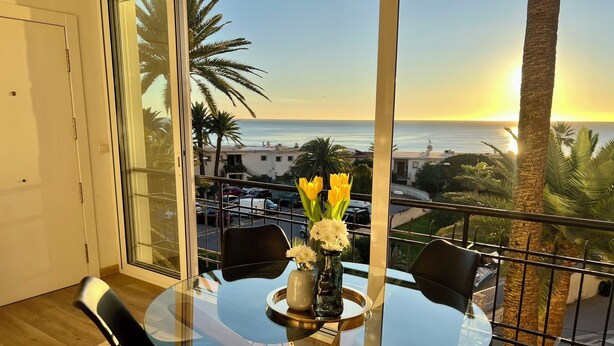 A balcony with a glass table and chairs, overlooking a sunset over the sea.