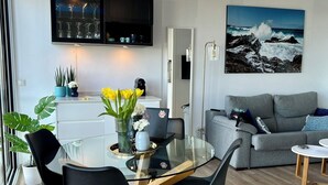 A modern living room with a glass dining table, black chairs, a grey sofa, and a large painting of a rocky coastline.