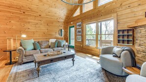 L’intérieur chaleureux d’un chalet en bois, avec un plafond en bois, une grande fenêtre, une cheminée en pierre et un coin salon confortable comprenant un canapé et un fauteuil.