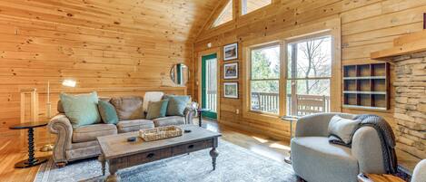 A cozy log cabin interior with a wooden ceiling, a large window, a stone fireplace, and a comfortable seating area with a sofa and armchair.