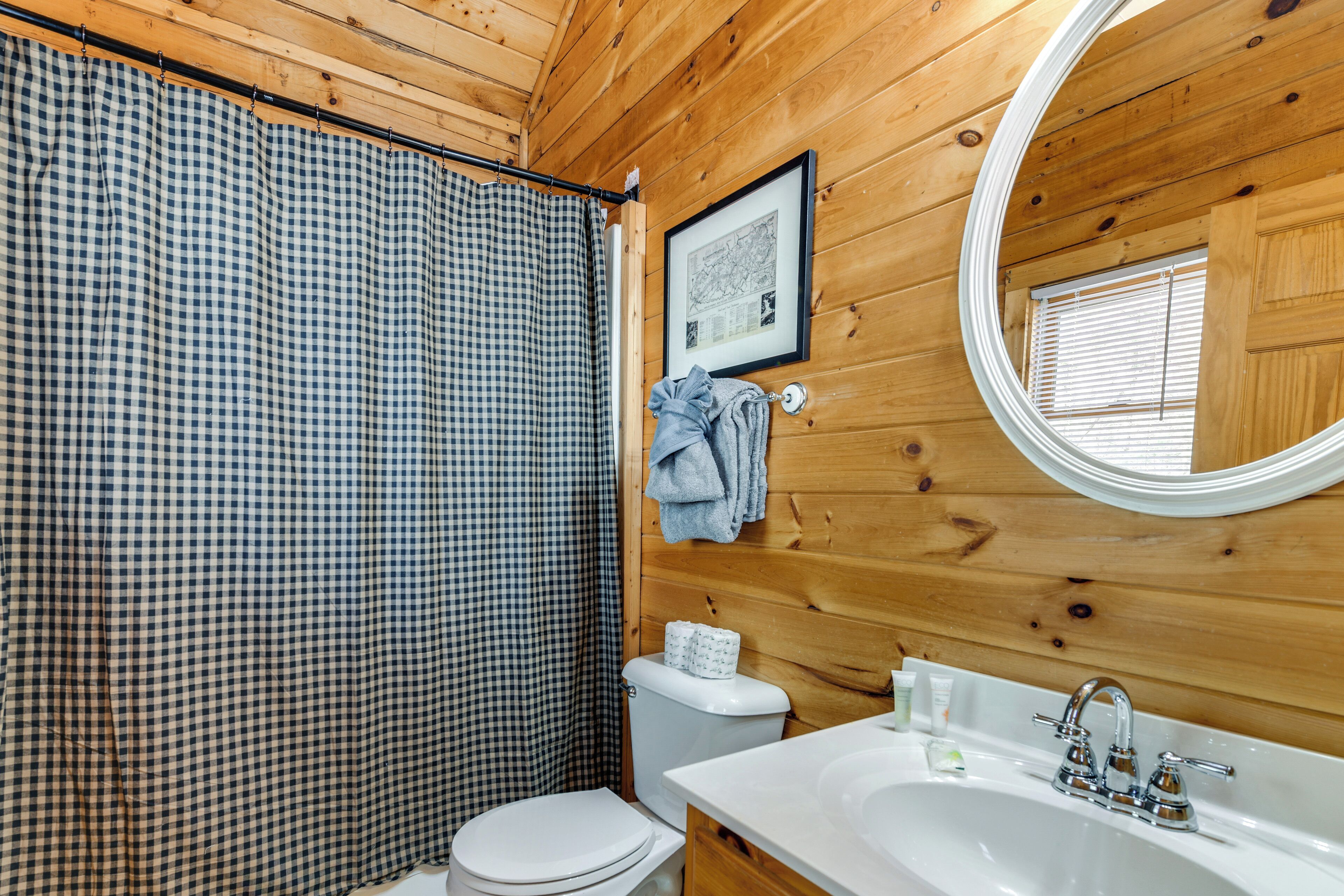 Baño de cabaña de madera con cortina de ducha a cuadros, un inodoro, un lavabo con espejo y un mapa enmarcado en la pared.