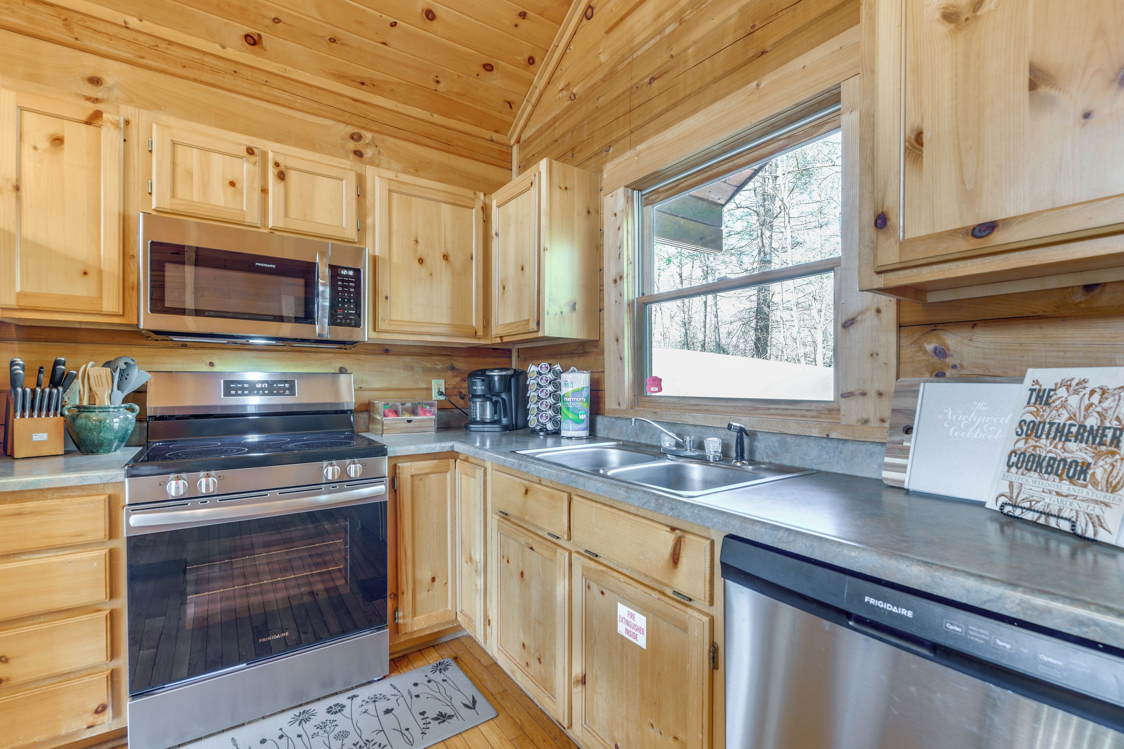 A modern kitchen with wooden cabinets, stainless steel appliances, and a large window providing a view of the outdoors.