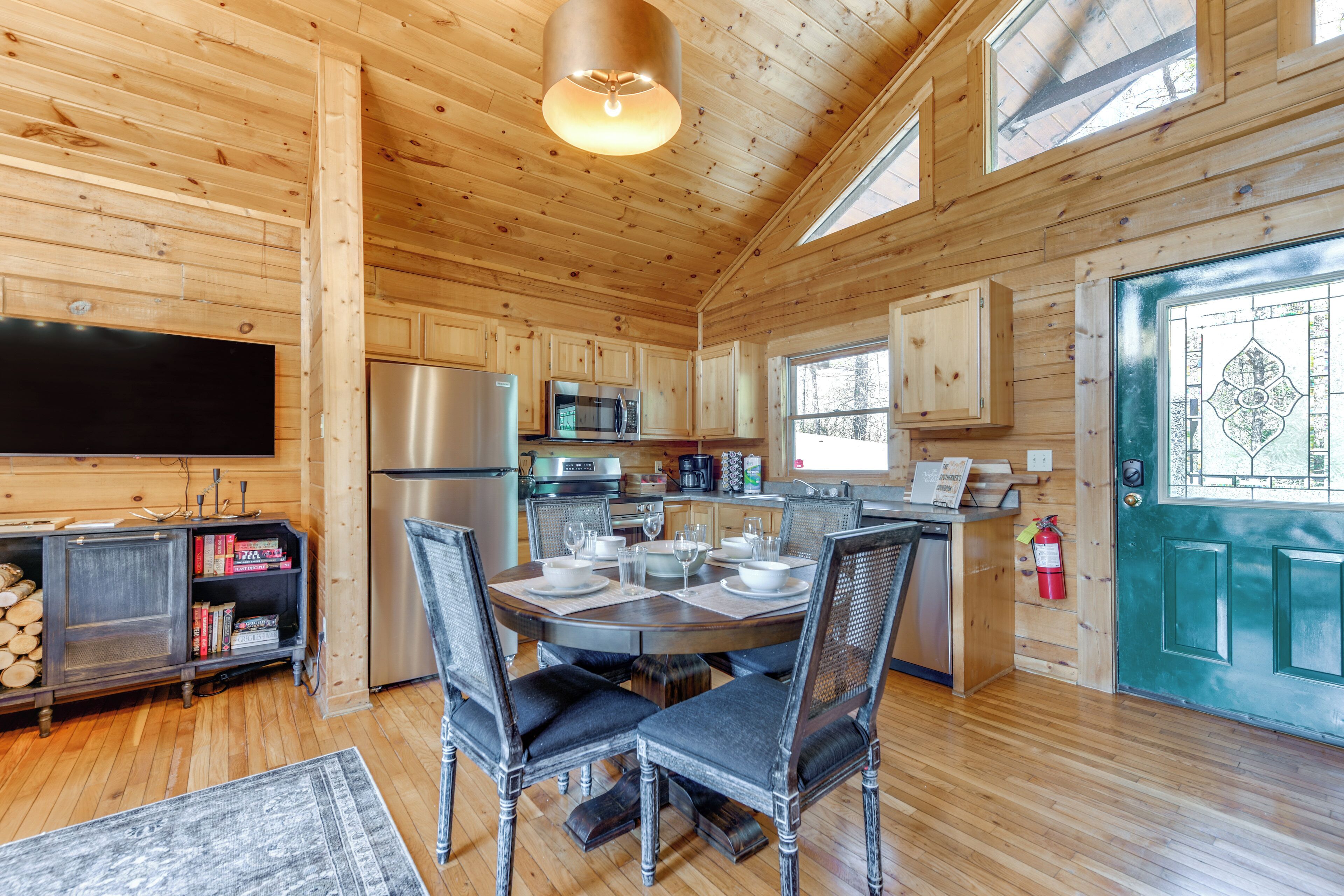 A wooden cabin interior with a dining table set for four, kitchen appliances, and a television mounted on the wall.