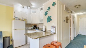 Une cuisine compacte avec des meubles blancs, un comptoir en granit et un petit îlot central. On y trouve deux tabourets orange et une poubelle.