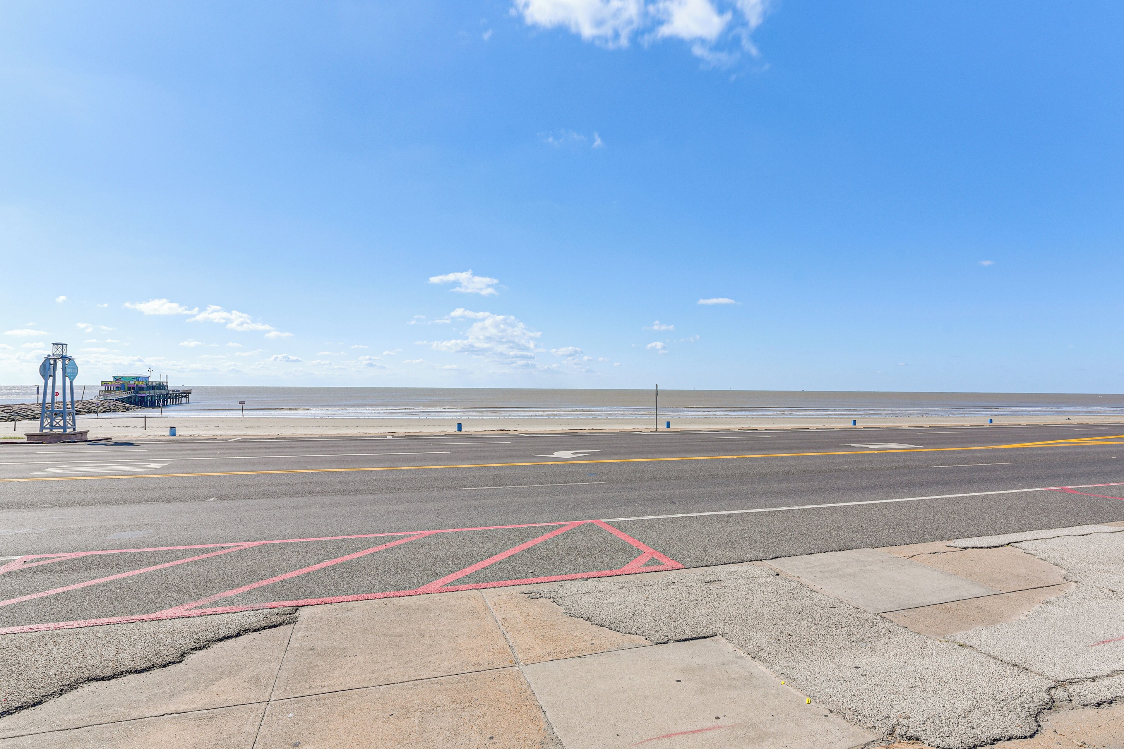 A coastal road with a clear sky, a beach, and a pier structure in the background.