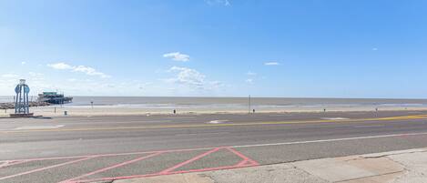A coastal road with a clear sky, a beach, and a pier structure in the background.