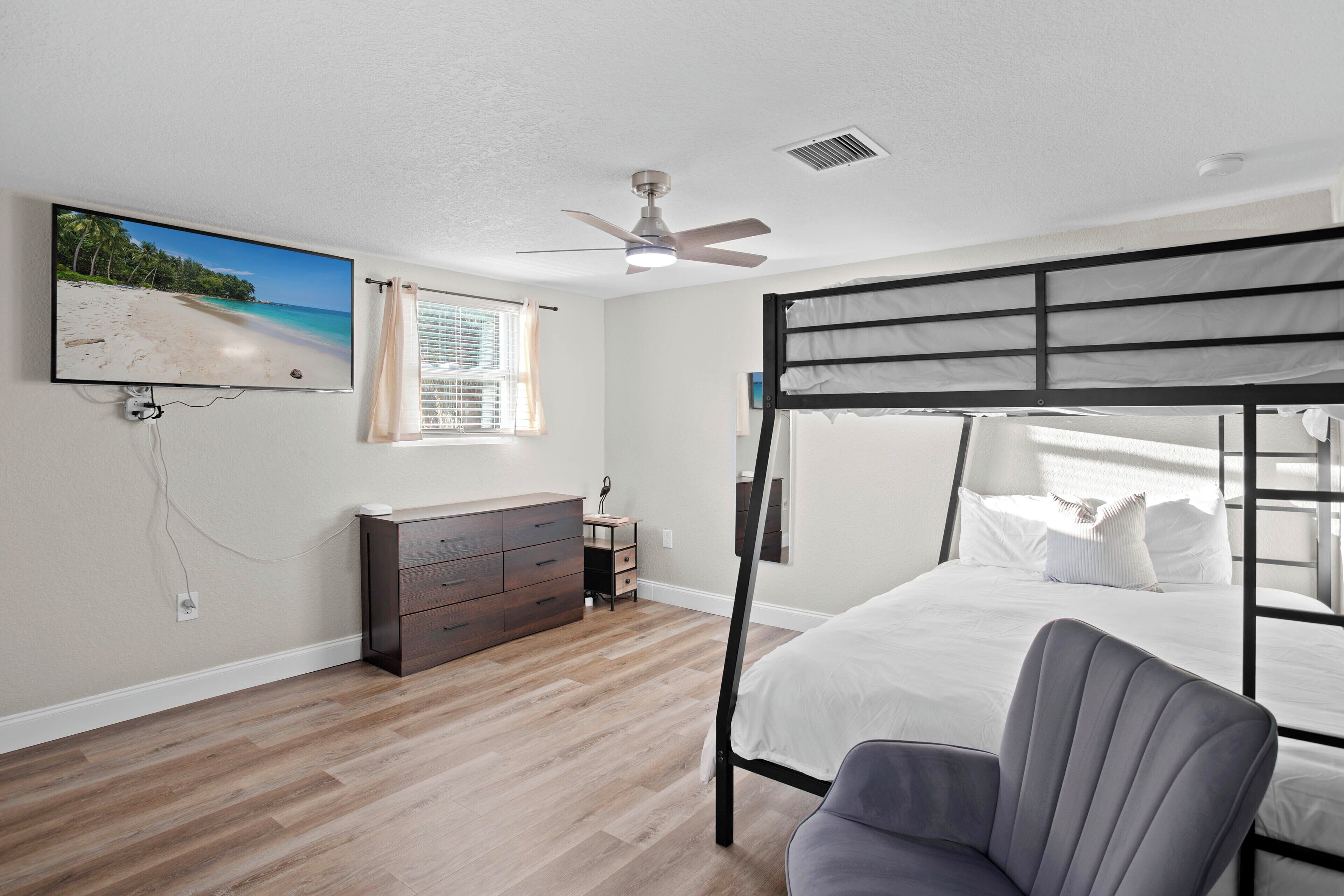 Une chambre avec un lit superposé, un ventilateur de plafond, un téléviseur fixé au mur, une commode et une fenêtre avec des rideaux.
