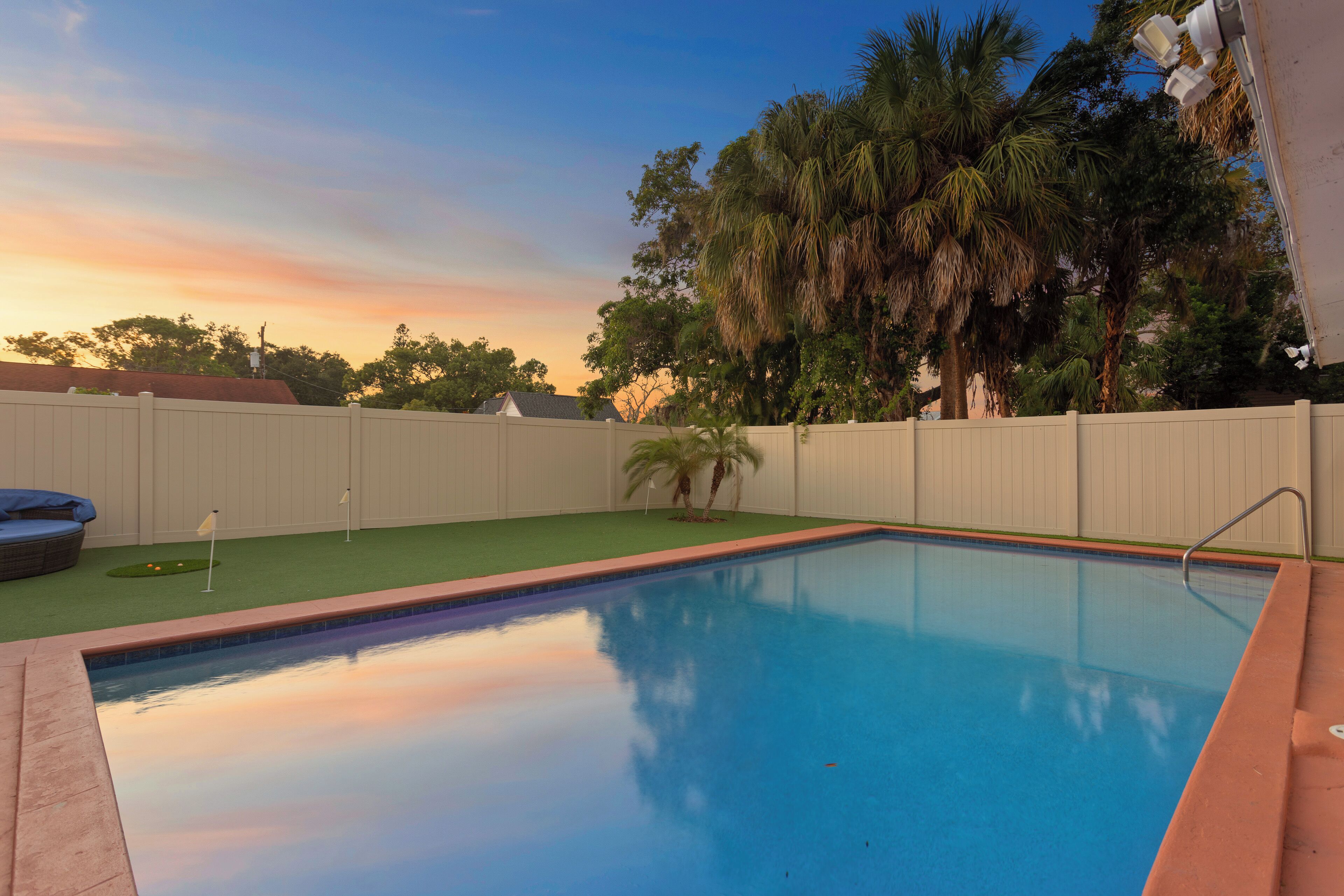 A rectangular swimming pool with a wooden deck edge, surrounded by a white fence and palm trees.