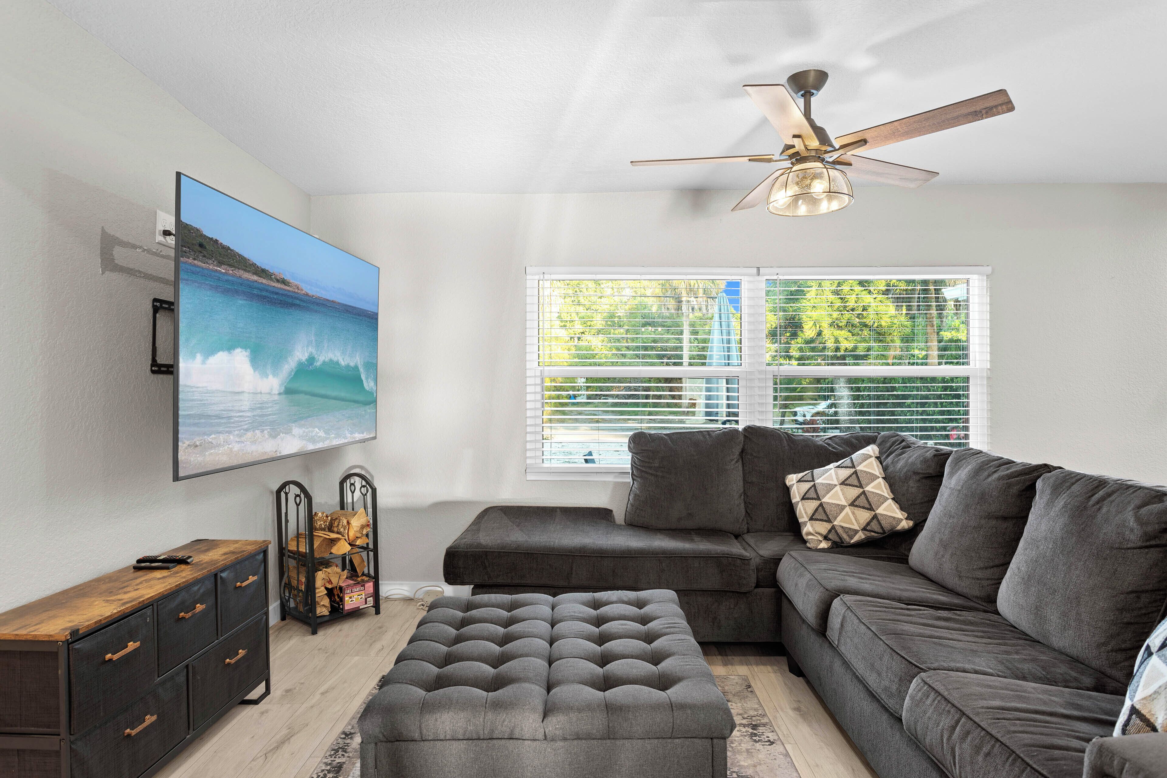 A living room with a large flat-screen TV, a ceiling fan, a sectional sofa, and a window with blinds.