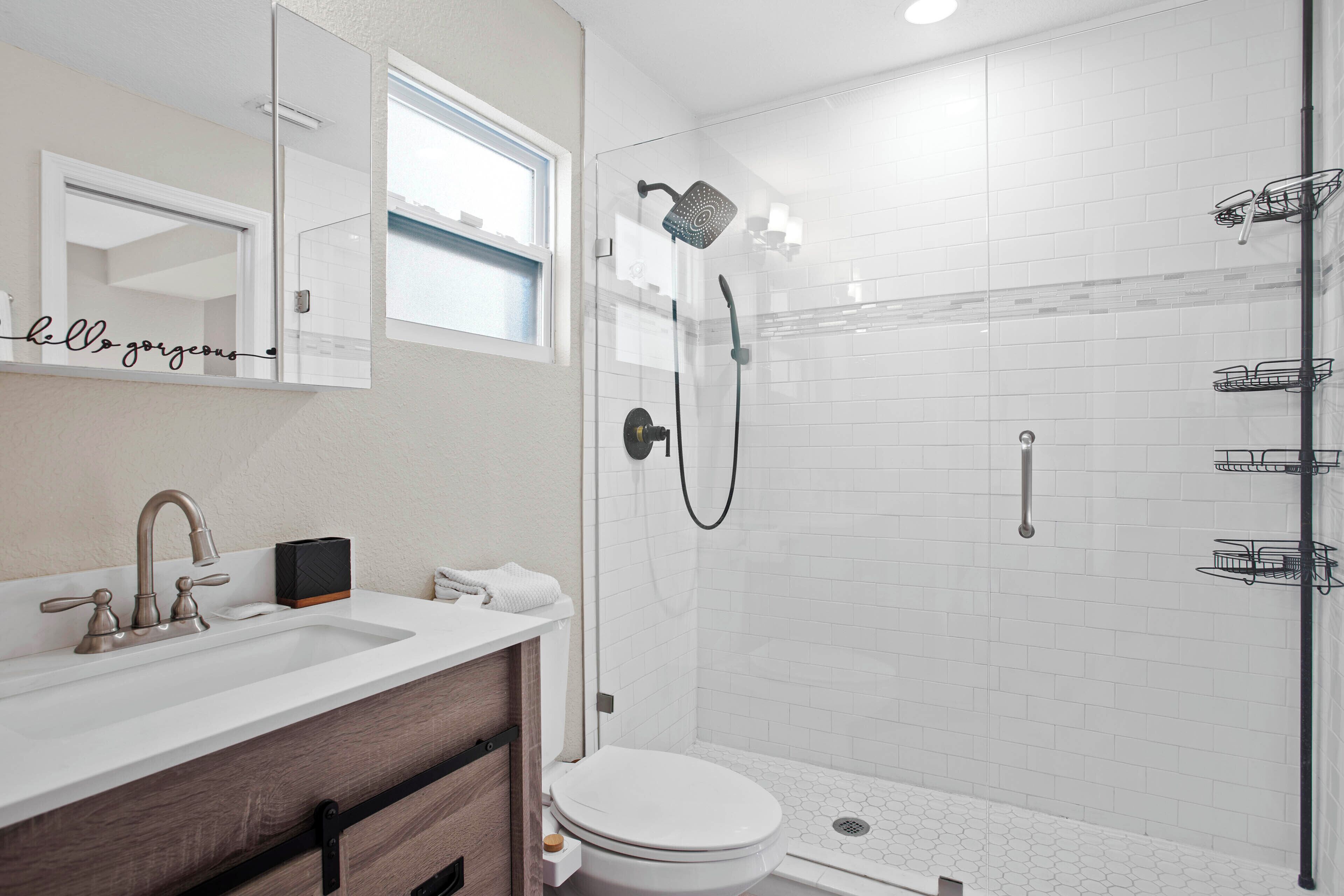 A modern bathroom with a white sink, vanity, and toilet. It includes a glass-enclosed shower with a handheld showerhead and a handheld showerhead with a rain showerhead.