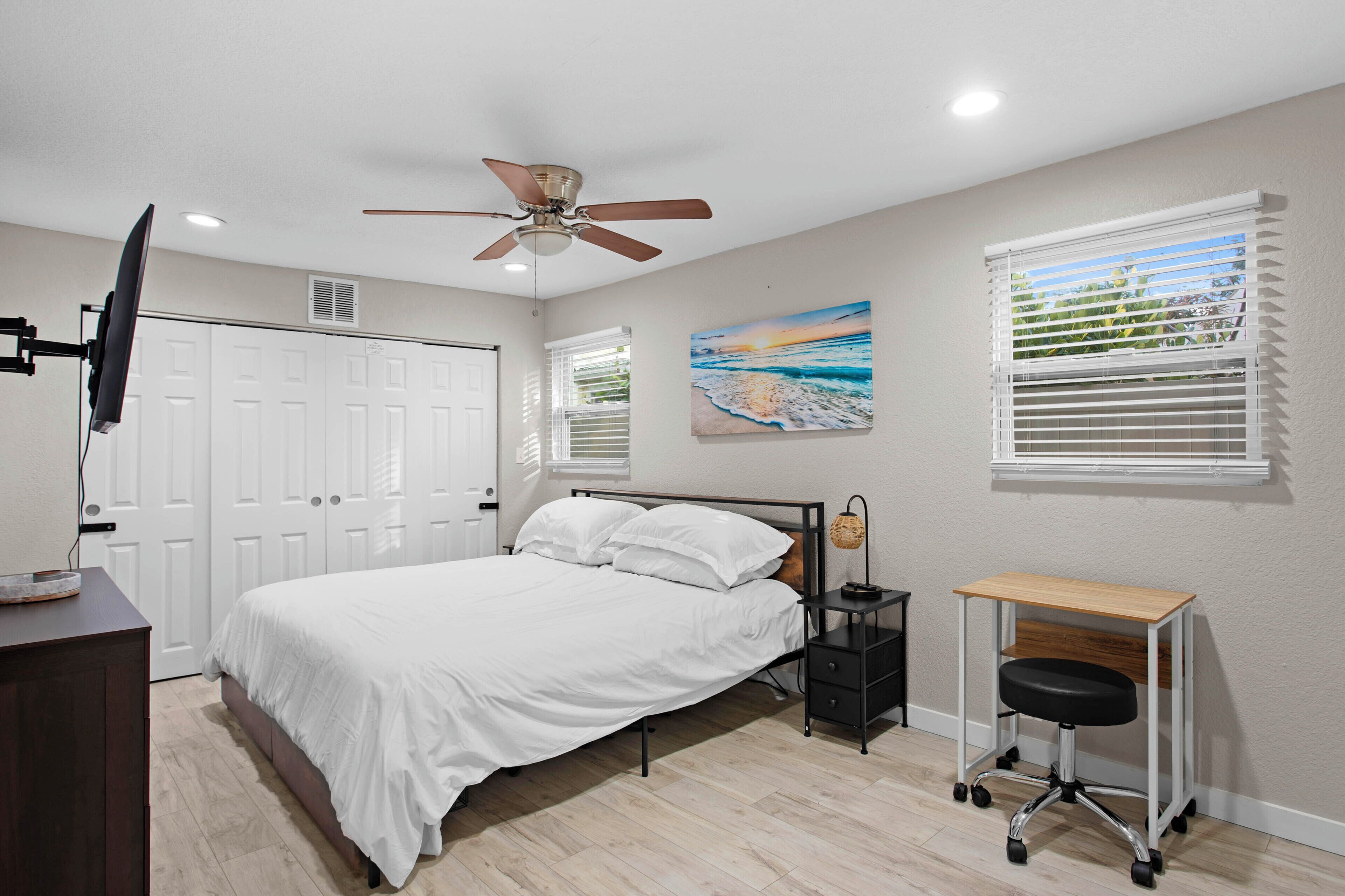 A bedroom with a bed, a desk, a chair, a ceiling fan, and a painting on the wall.