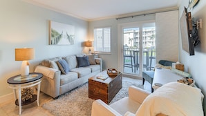 A modern living room with a sofa, a coffee table, and a side table. There is a window with blinds, a lamp, and a view of the outside. - Beachfront Retreat w/ Pool Access in Gulf Shores! (Gulf Shores)