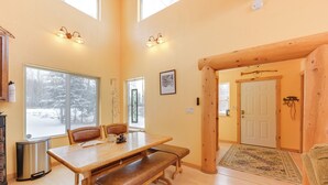 A spacious dining area with a wooden table and benches, large windows with a snowy view, and a wooden door leading to another room. - 5 Mi to Kasilof River: Forest-view Retreat w/ Deck (Kasilof)