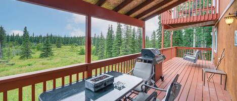 A covered outdoor deck with a table and chairs, a grill, and a view of a forest.