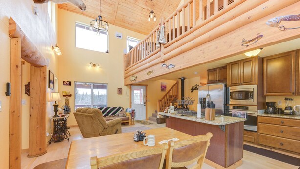 A spacious living area with a wooden dining table, two chairs, a sofa, and a fireplace. A kitchen with wooden cabinets and appliances is visible in the background. - 5 Mi to Kasilof River: Forest-view Retreat w/ Deck (Kasilof)