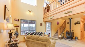 A cozy living room with a large window, a wooden staircase, and a wood-burning stove. - 5 Mi to Kasilof River: Forest-view Retreat w/ Deck (Kasilof)