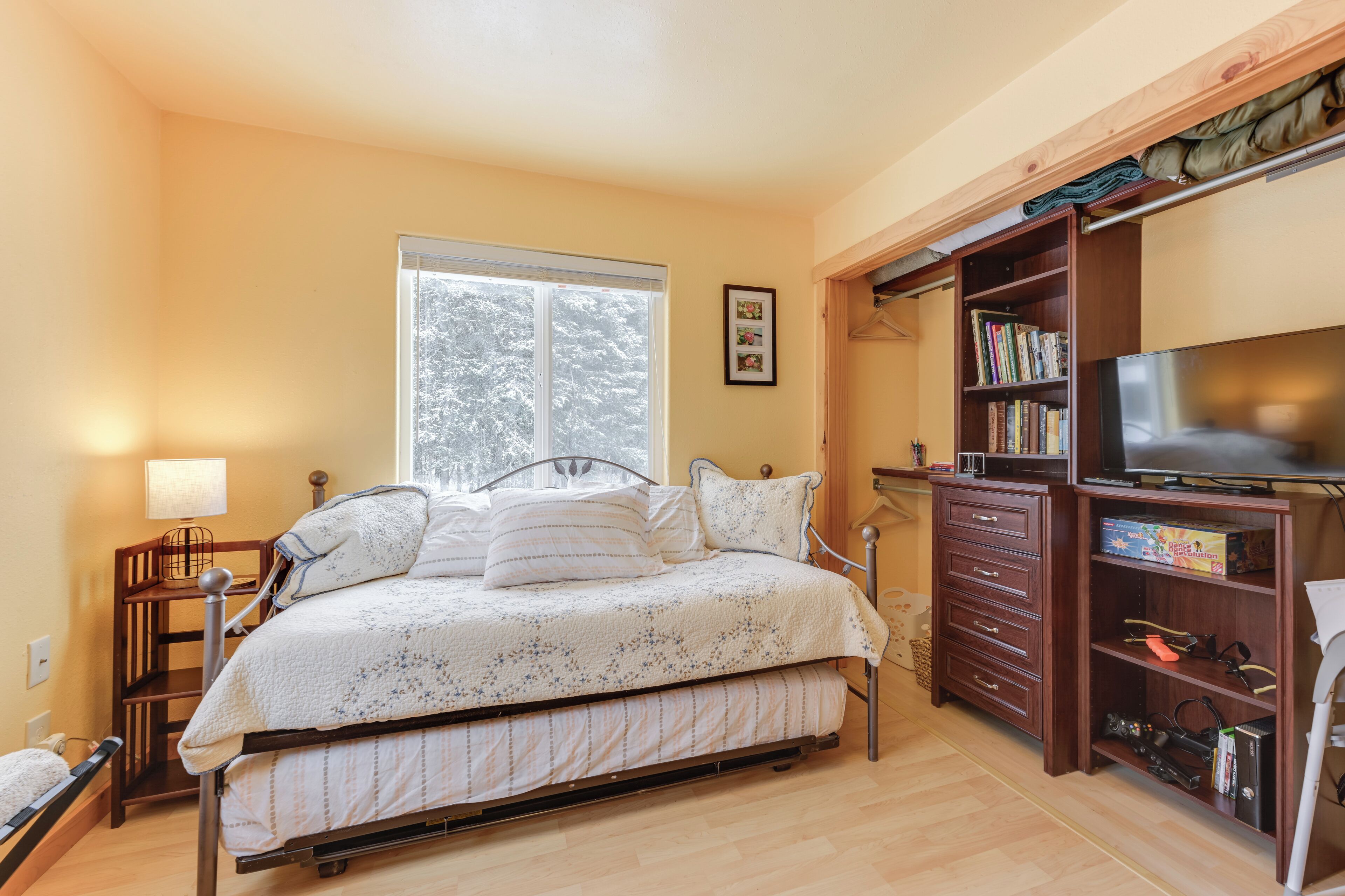 Un dormitorio con cama, televisión, estantería y ventana con vista a árboles.