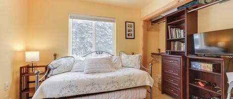 Un dormitorio con cama, televisión, estantería y ventana con vista a árboles.