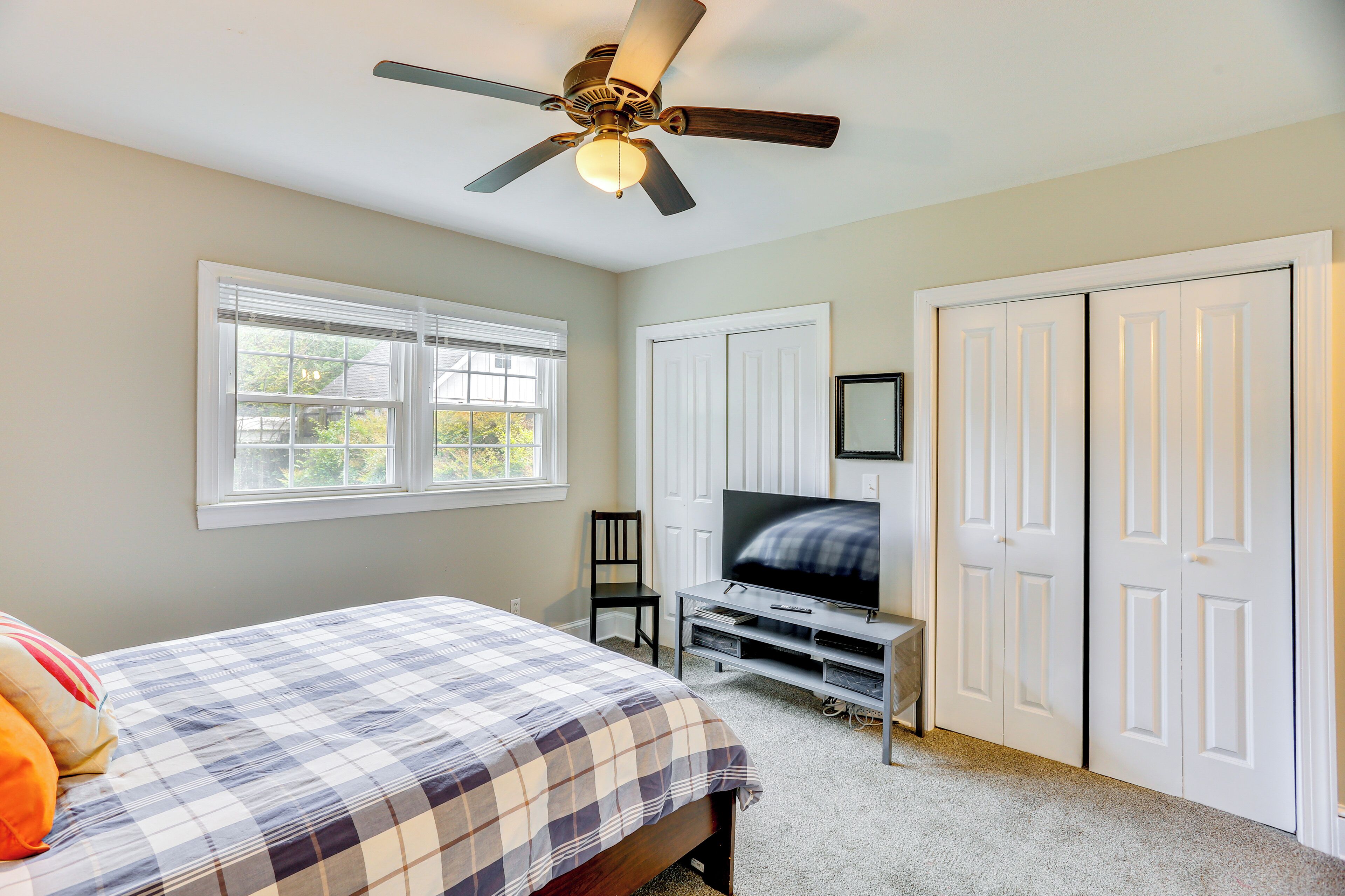 A bedroom with a bed, a TV on a stand, a ceiling fan, and a window.