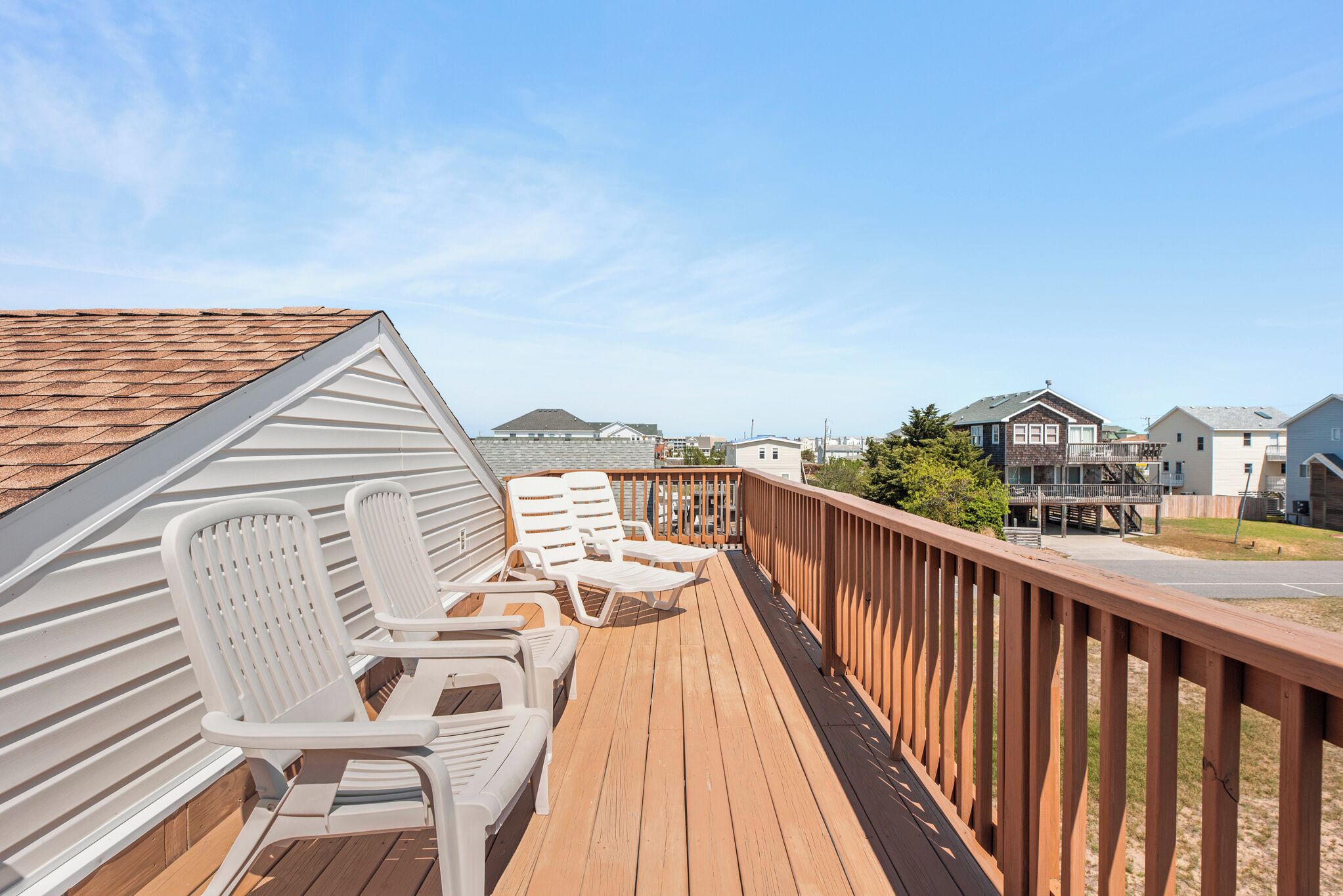 A wooden deck with white lounge chairs, a wooden railing, and a view of residential buildings in the background.