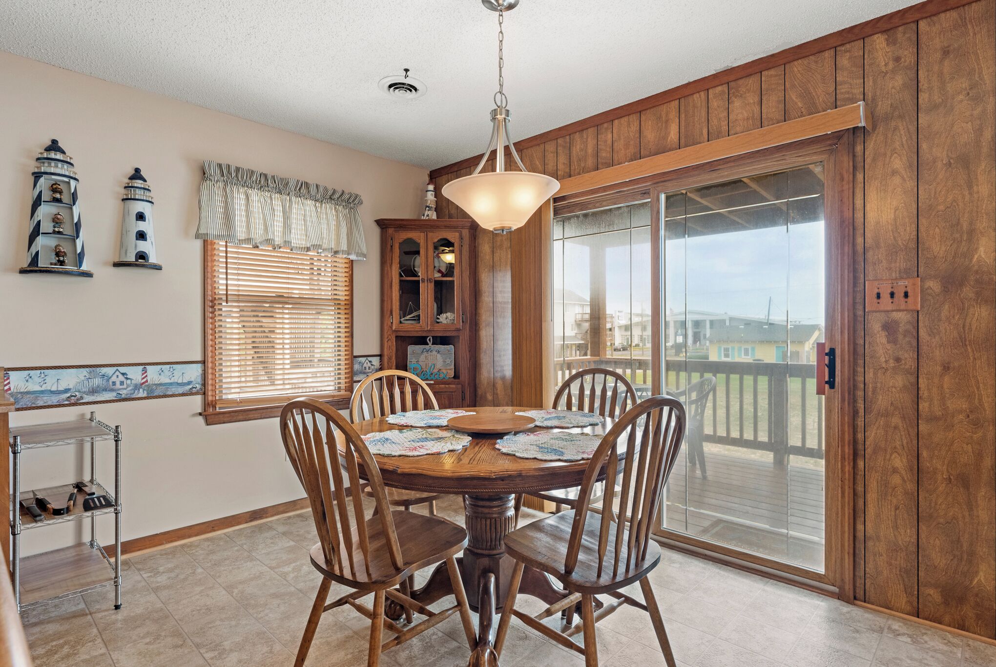 A dining area with a wooden table and chairs, a chandelier, a window with blinds, and a view of a deck.