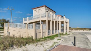 Egy többemeletes épület erkélyekkel, a háttérben egy homokos strand, és egy fából készült sétaút vezet oda.