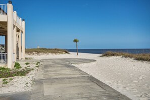 Ein Betonweg führt zu einem Strand mit einer Palme und einem Gebäude mit einem Balkon.