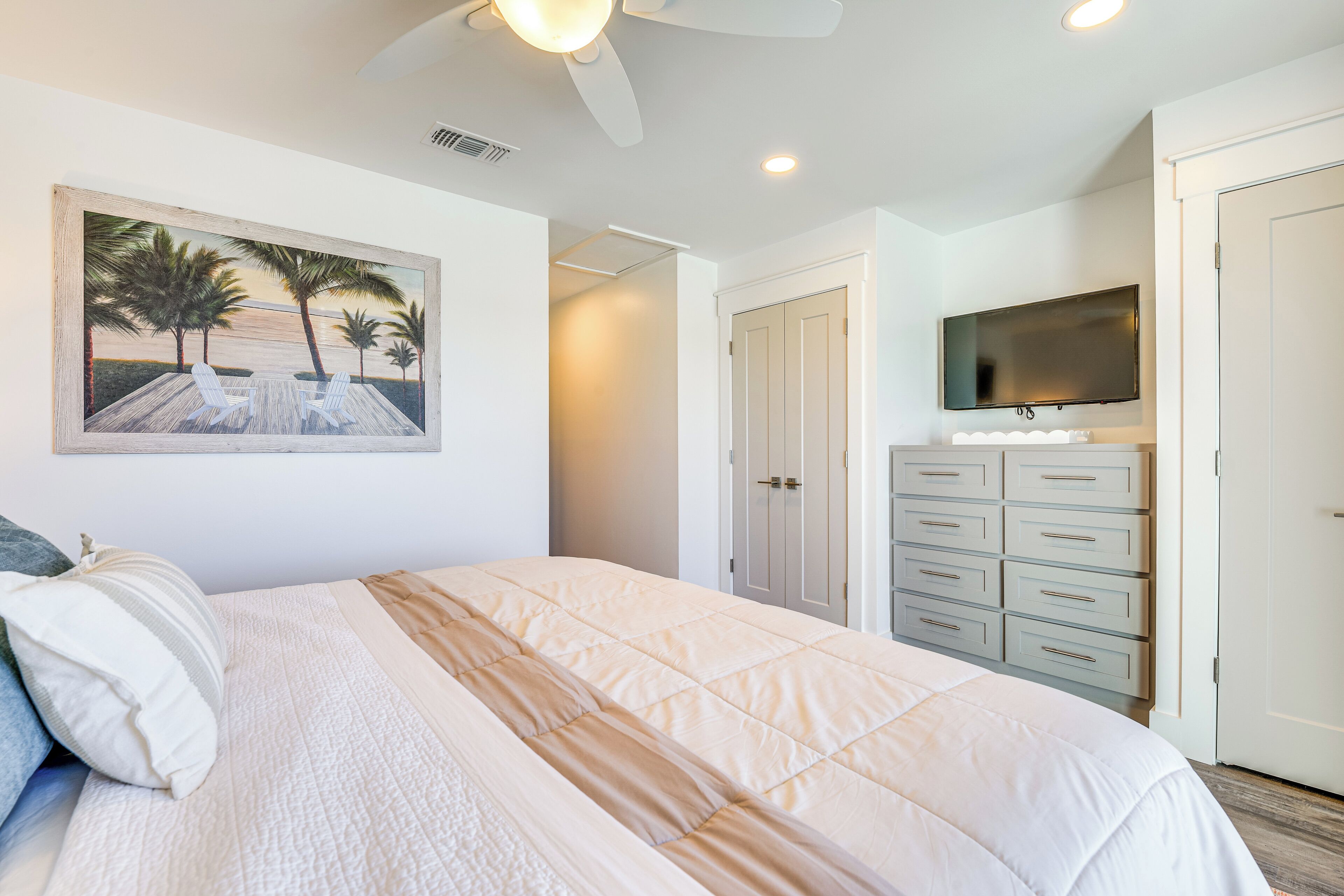 A bedroom with a bed, a painting of a beach scene, a ceiling fan, a TV, and a dresser.