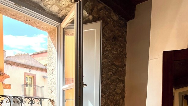 Una habitación con pared de piedra, ventana con vista a un balcón y una silla blanca con una toalla roja.