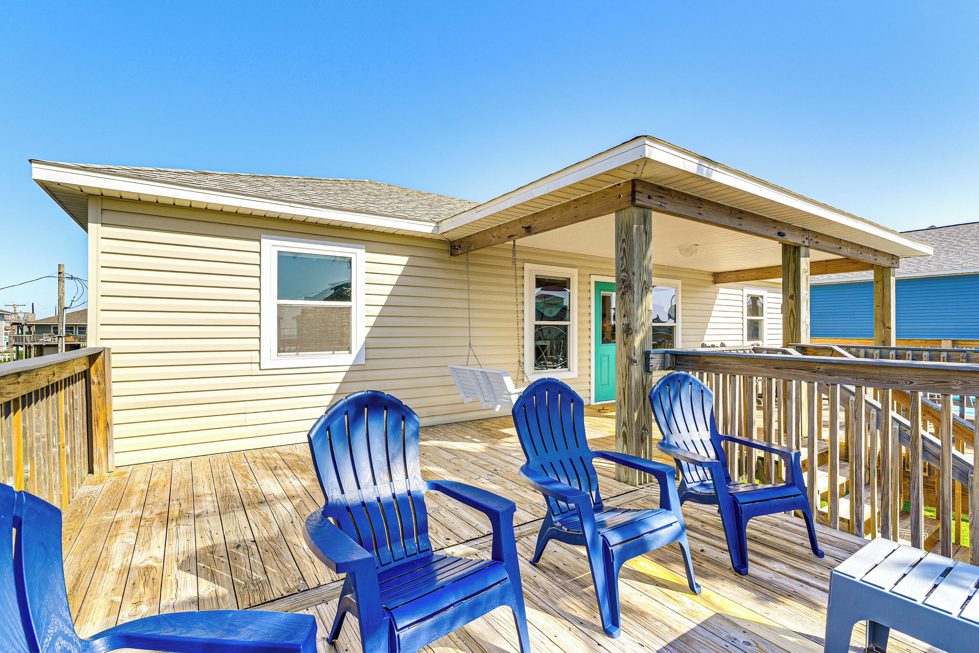 A wooden deck with blue Adirondack chairs, a covered porch, and a beige building with a green door.