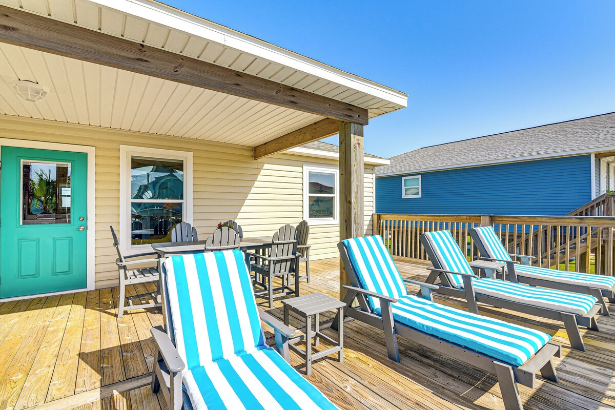 A wooden deck with lounge chairs and a table, a turquoise door, and a blue building in the background.