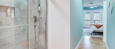 Un baño moderno con cabina de ducha de vidrio, baldosas de mármol y suelo de madera que conduce a un dormitorio con cama y ventilador de techo.