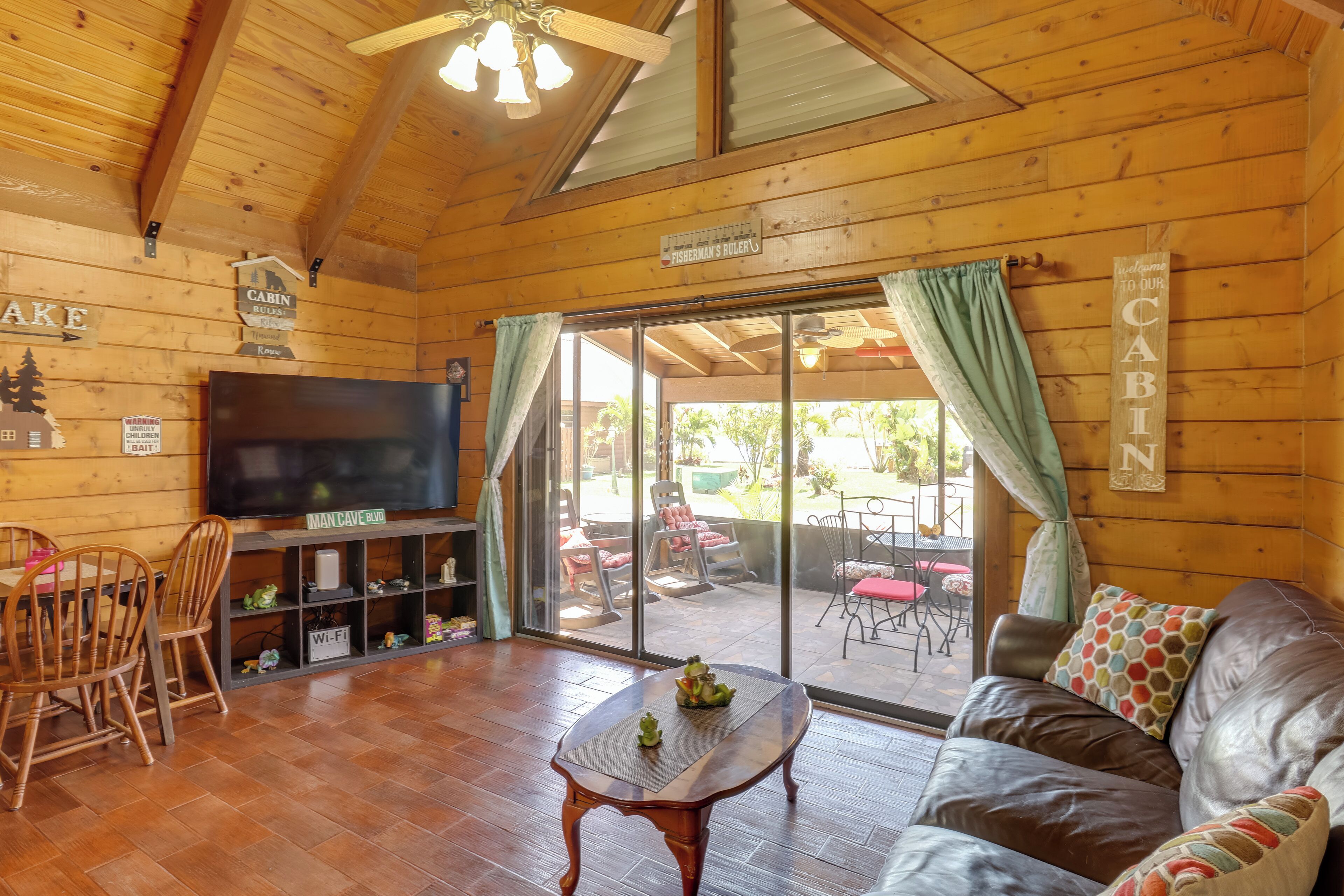 Interior de una cabaña con techo de madera, un televisor, una mesa con figuras de ranas, un sofá con almohadones estampados y un ventanal corredero que da a un patio exterior.