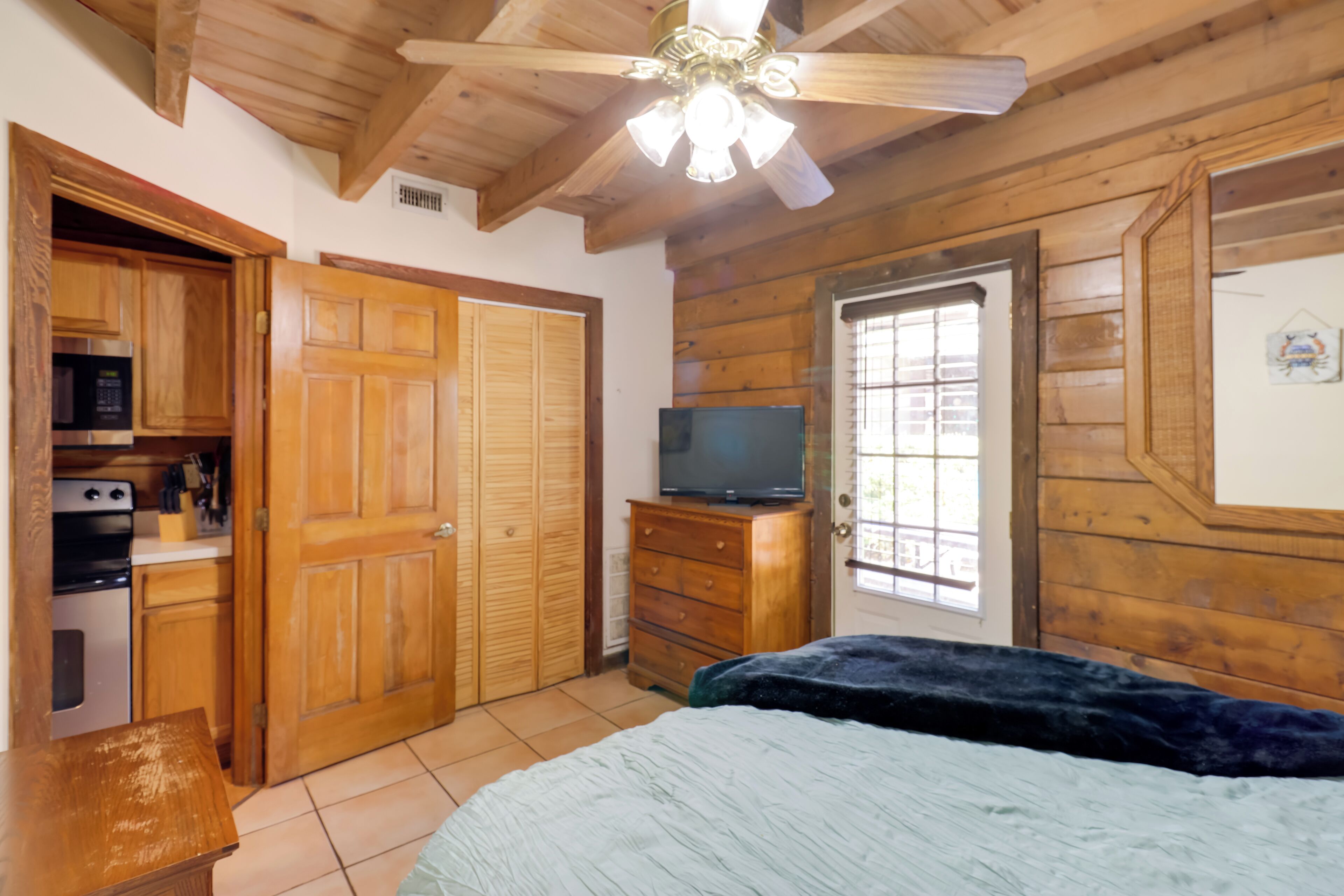 A bedroom with wooden walls, a ceiling fan, a bed, a dresser, a television, and a window.