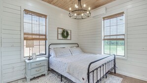 A bedroom with a bed, a nightstand, a chandelier, and two windows with blinds. - Contrell Cottage On Farm w/ Peaceful Views (Lena)