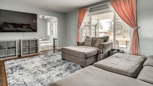 A modern living room with a sectional sofa, a flat-screen TV mounted on the wall, and a large window with curtains.