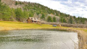 A cabin with a porch and a car parked nearby, surrounded by a lake and forested hills.