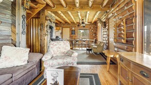 Ein Holzhaus-Interieur mit Holzbalkendecke, einem floralen Sessel, einem Sofa, einem Esstisch mit Stühlen und einem Holzschrank.